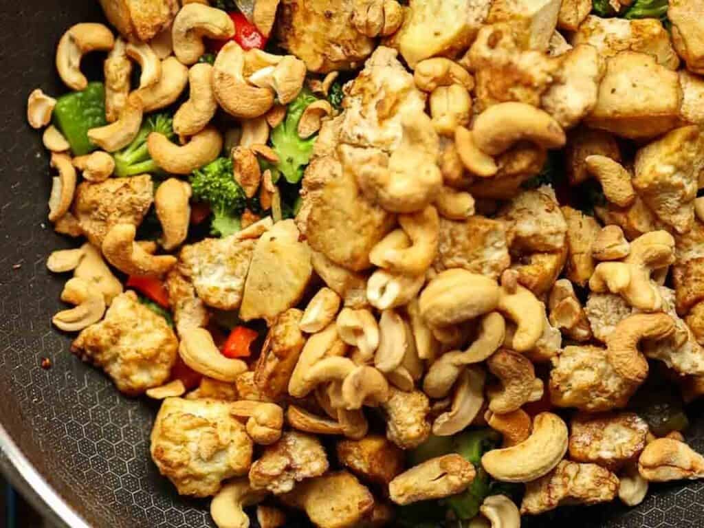 A pan filled with a colorful stir-fry featuring chunks of tofu, broccoli, red bell pepper, and a generous amount of cashew nuts. The ingredients are lightly sautéed, giving the dish a vibrant and appetizing appearance.